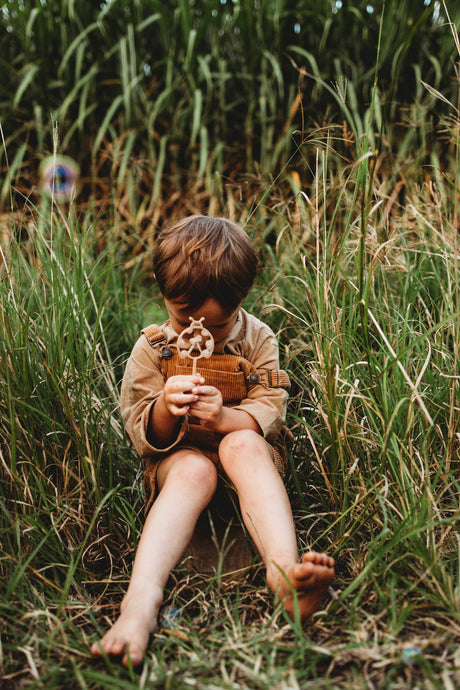 A young child sitting barefoot in tall green grass, holding a small brown leather pouch and a decorative wand.