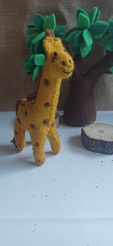 Yellow felt giraffe toy with brown spots standing next to a tree and log on a white surface.