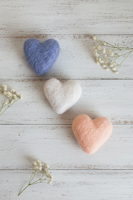 Three felted wool hearts in dusty blue, off-white, and soft peach, arranged on a textured beige linen surface with small dried flowers and a green eucalyptus sprig.
