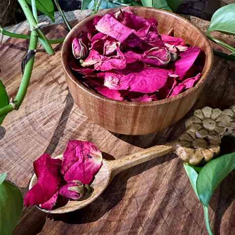 Wooden bowl and spoon filled with pink rose petals on a wooden surface.