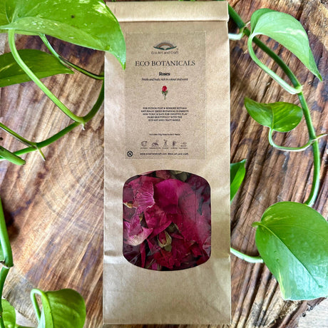 A kraft paper bag of Eco Botanicals Rose herbal tea with a clear window showing dried pink rose petals, styled on a rustic wooden surface with green pothos plant leaves.