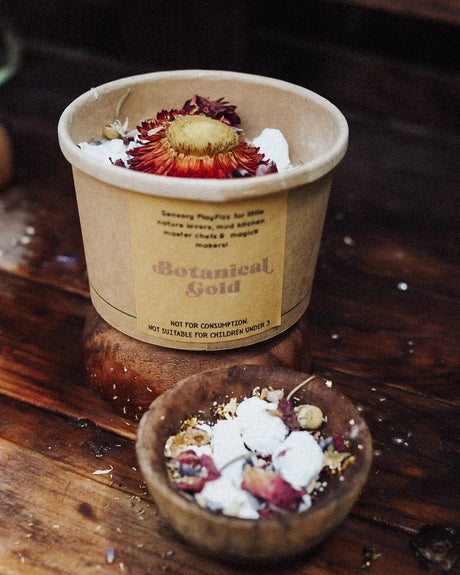 A small wooden bowl filled with white powder and dried botanical pieces, with a blurred container labeled 'Botanical Gold' in the background on a rustic wooden surface.