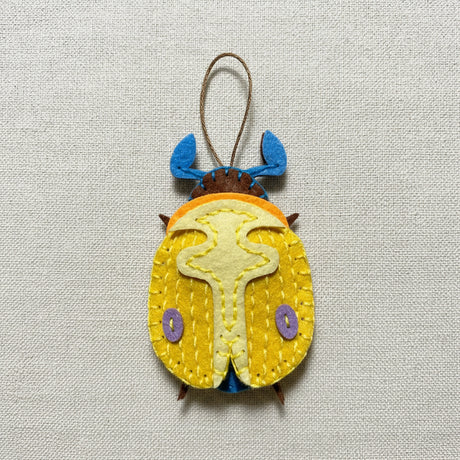 A handmade felt beetle with yellow wing cases, blue antennae, and a brown body, hanging by a string on a creme background.