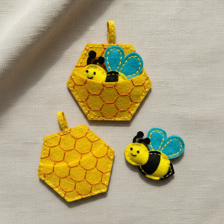 Three yellow felt bee and honeycomb-themed ornaments on a plain white background, including two hexagonal honeycomb shapes and one smiling bee figure.