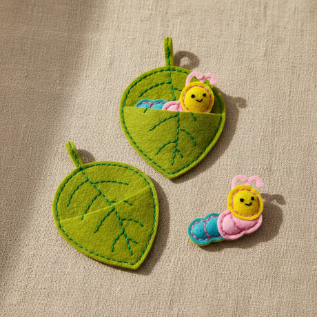 Two green felt leaf-shaped hanging pouches with embroidered leaf veins, shown on a white background. One pouch contains a small plush caterpillar toy, and a second matching caterpillar is placed beside it.