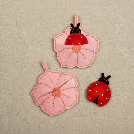 Three pink flower-shaped felt items with ladybugs. Two are arranged as open flowers and one is closed, showing a red ladybug with white polka dots and black antennae.