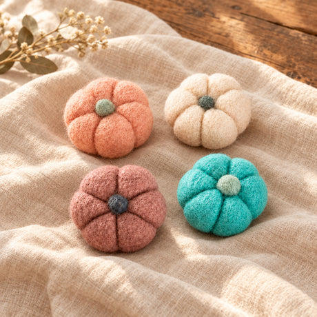 Four hand-felted mini pumpkins in peach, light pink, terracotta, and turquoise, arranged on a background of brown kraft paper.