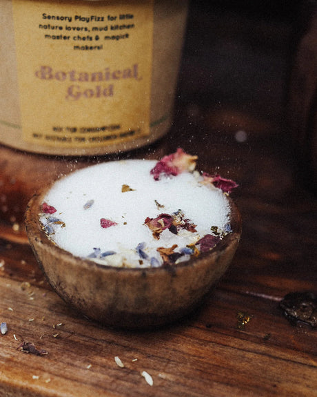 A small wooden bowl filled with white powder and dried botanical pieces, with a blurred container labeled 'Botanical Gold' in the background on a rustic wooden surface.
