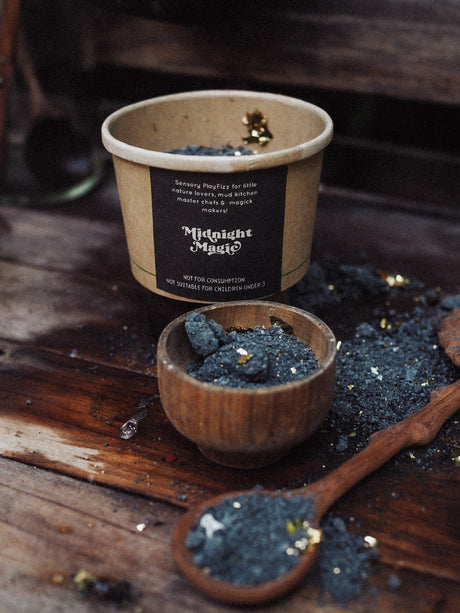 A kraft paper cup labeled 'Midnight Magic' filled with dark moss, shown on a rustic wooden surface with a small wooden bowl, a wooden spoon, and scattered moss pieces.