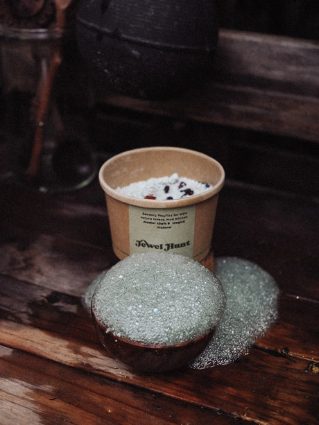 A bowl filled with pale green sparkling bath salts sits on a rustic wooden surface, with a kraft paper cup behind it containing white bath salts and dark botanical pieces. The cup has a label reading 'Jewel Hunt'.