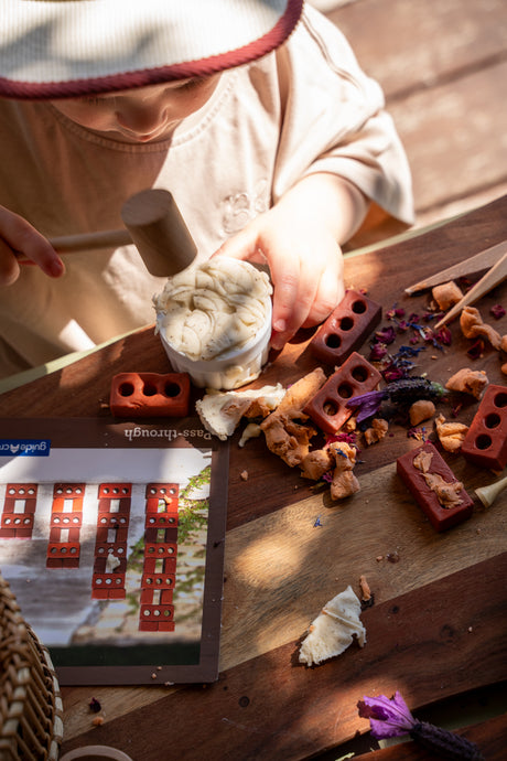 A child wearing a cream-colored outfit and a white hat with a red trim is actively playing with a wooden toy set on a rustic wooden table. The toy set includes red brick-like pieces, a small hammer, and a white clay-like block being tapped.