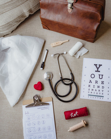 A styled flat-lay of a vintage brown leather medical kit bag surrounded by medical-themed props on a beige fabric surface, including a stethoscope, prescription pad, eye chart, thermometer, bandage, and small red heart.