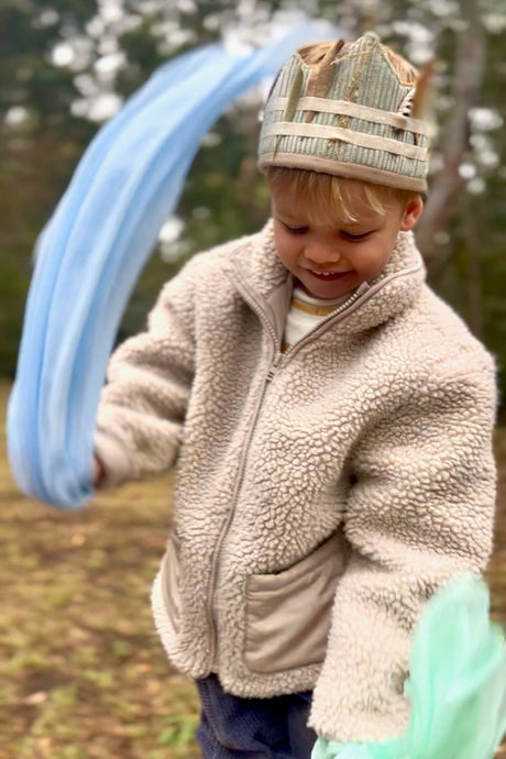 Child in a beige fleece jacket holding blue and green balloons outdoors.