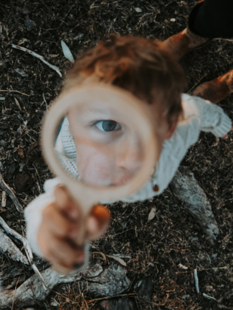 A close-up lifestyle shot of a young child holding a magnifying glass, looking through it at the camera on a forest floor background.