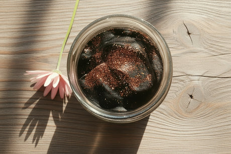 A clear glass jar filled with black playdough and glitter on a wooden surface with a pink flower beside it and warm sunlight casting shadows.