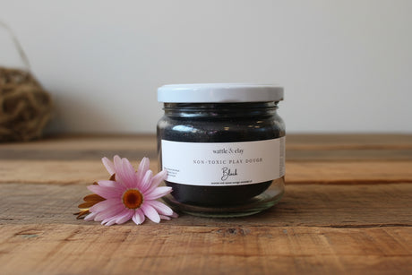 A glass jar candle filled with charcoal-colored play dough, featuring a white lid and a white label that reads 'wattle & clay' A pink flower is placed next to the jar on a light wooden surface with a wooden background.
