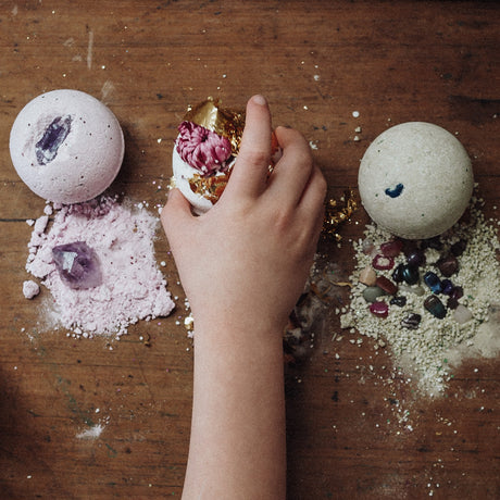 A child's hand peels back a decorative fizz egg on a wooden table, with two unopened fizz balls and scattered gemstones and powder around them.