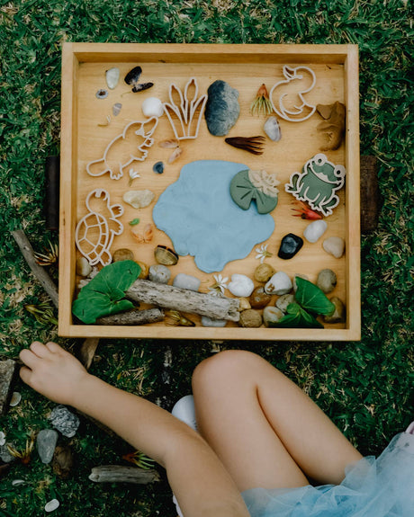 Wooden tray with nature-themed items on grass, child's legs visible