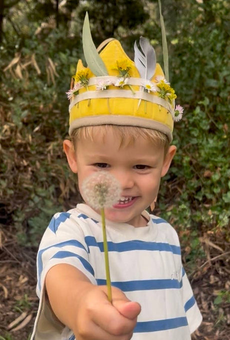 Child wearing a yellow hat with plant decorations, holding a dandelion in a natural setting.