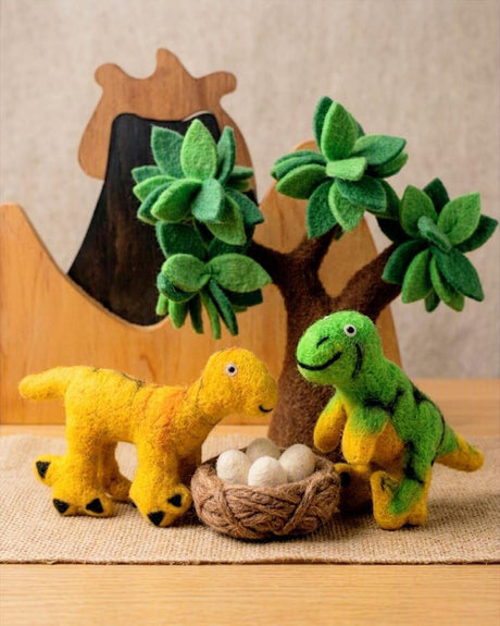 Handmade felt dinosaur toys and a small felt tree with green leaves, arranged on a burlap mat with a wooden background.