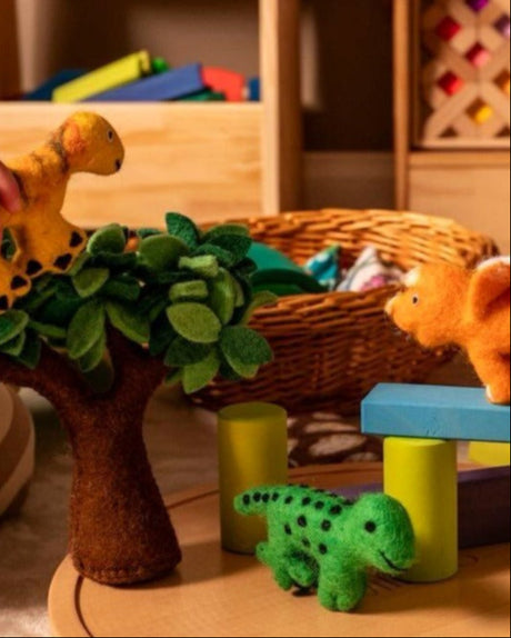 A cozy indoor play scene with children's wooden and felt dinosaur toys arranged on a round wooden base. Hands are visible holding the toys, and shelves of colorful blocks and educational toys fill the background.