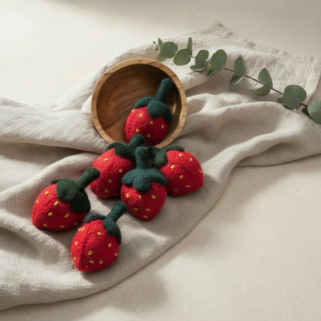 Five red felt strawberry ornaments with dark green stems and yellow seed details are arranged on a woven mat, with one nestled in a wooden bowl and two near decorative butterflies. Red dried flowers and a wood-paneled background complete the styled scene.