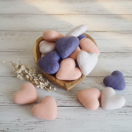A heart-shaped wooden bowl filled with soft felted wool hearts in pastel pink, lavender purple, and white, styled on a neutral beige fabric background with a few hearts placed outside the bowl and dried floral decor to the side.