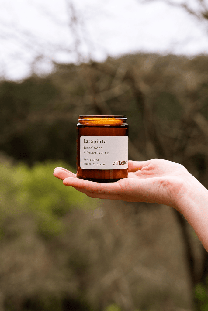 Hand holding a candle jar labeled 'Larapinta' against a blurred natural background