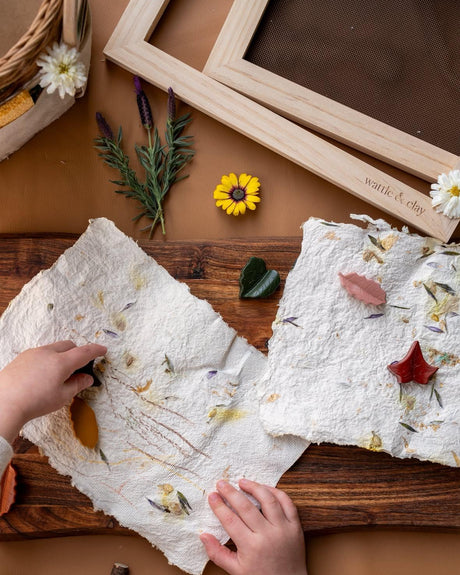 A child's hands press dried flowers and leaves into textured handmade paper laid on a wooden table, with two wooden frames and small botanical props around it.