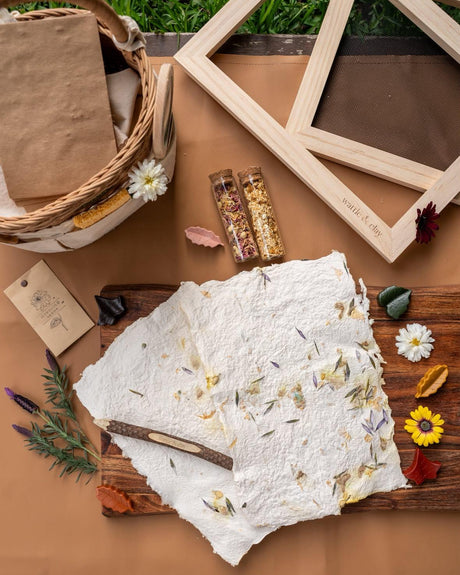 A styled flat-lay of a handmade paper craft kit on a brown surface, featuring a white textured sheet with dried botanicals, a wooden frame, a small basket with brown paper, and two small glass vials containing dried flower and seed mixtures.