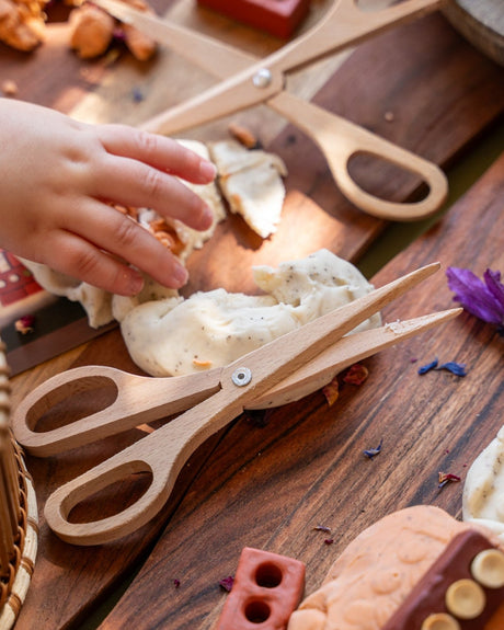 A close-up of a child's hand playing with wooden toy scissors and play dough on a rustic wooden table, surrounded by red brick-like blocks and craft materials.