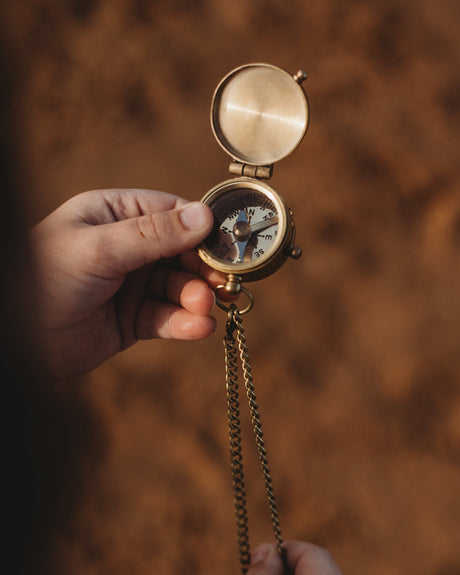 A vintage brass compass with an open lid and chain, held by a child against a warm, blurred brown background.