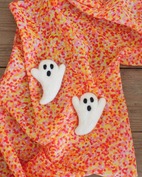 Two white felt ghost-shaped accessories with black embroidered eyes and mouths, placed on a bright orange, pink, and yellow patterned fabric background.