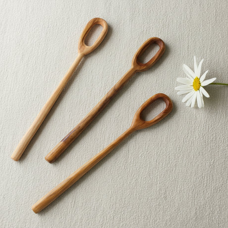 Three teak stirrers arranged diagonally on a light gray textured surface next to a white daisy flower.