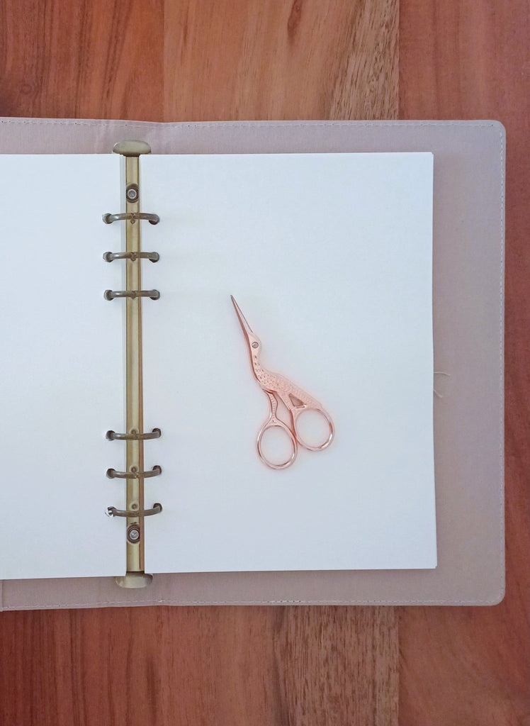 Rose gold scissors on a white sheet of paper inside a notebook with a metal binding on a wooden surface.