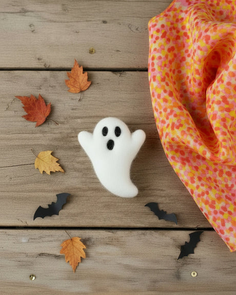 A small white felt ghost accessory with black embroidered eyes and mouth, resting on a crumpled piece of orange, pink, and yellow patterned paper against a black background.