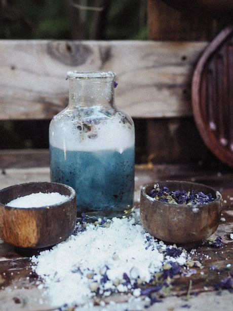 A rustic glass bottle filled with blue bath salts sits on a wooden surface, surrounded by bowls of white bath salts and dried purple flowers, with a blurred wooden background.