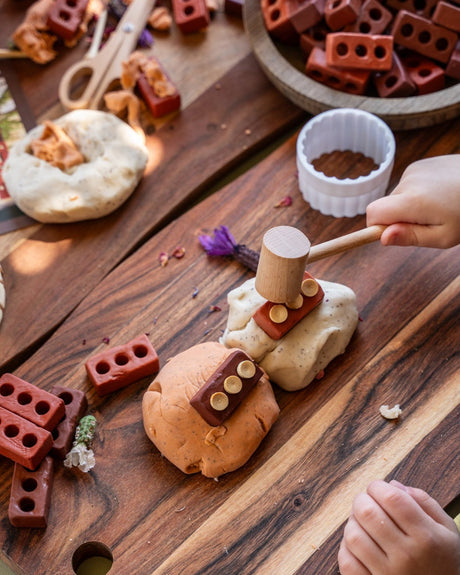 A child's hands are shown playing with a wooden mallet and red blocks on a a rustic wooden board with playdough.