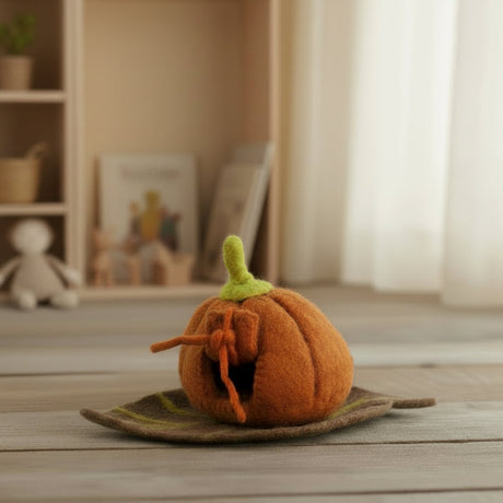 A small orange felt pumpkin decoration with a green stem sits on a leaf-shaped tray on a light wooden floor in a softly lit room with toys and shelves in the background.